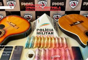 Material apreendido pela Polícia Militar durante a Operação Sentinela em Nepomuceno, incluindo instrumentos musicais, celulares, dinheiro e porção de droga, expostos em mesa, além de viaturas da PM estacionadas em via urbana.